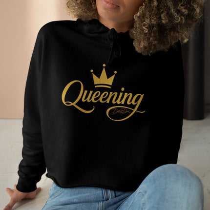 Woman wearing a black hoodie with 'Queening' printed in gold, sitting on the floor.