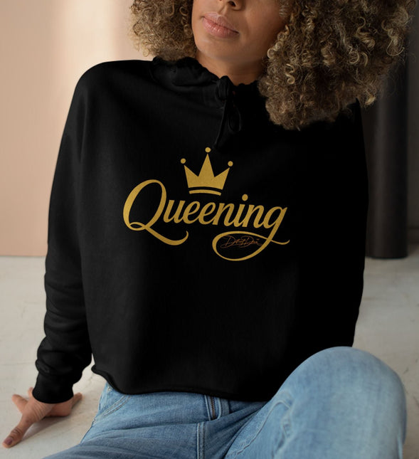 Woman wearing a black hoodie with 'Queening' printed in gold, sitting on the floor.