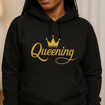 Person wearing a black hoodie with 'Queening' printed in gold on a beige background