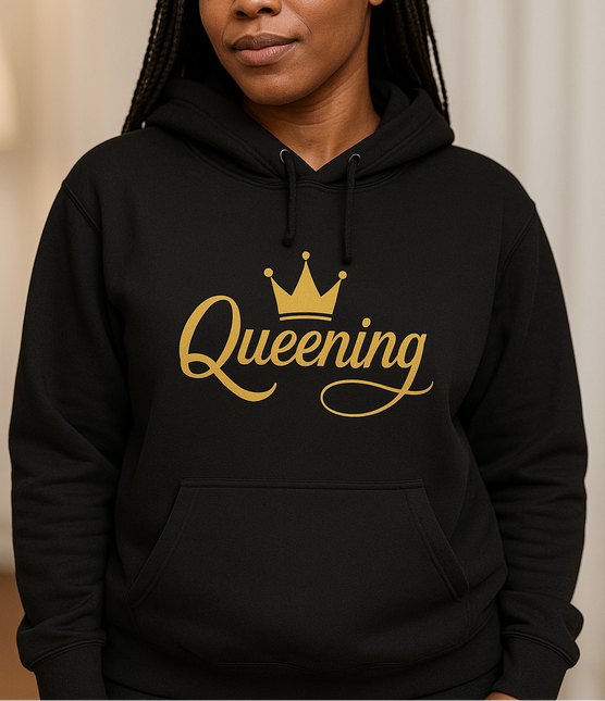 Person wearing a black hoodie with 'Queening' printed in gold on a beige background